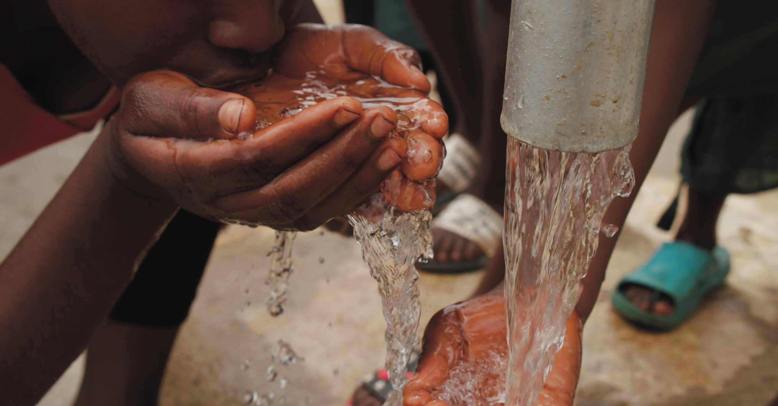 Close-up of hands and face drinking fresh water from an outdoor tap, emphasizing refreshment and necessity.